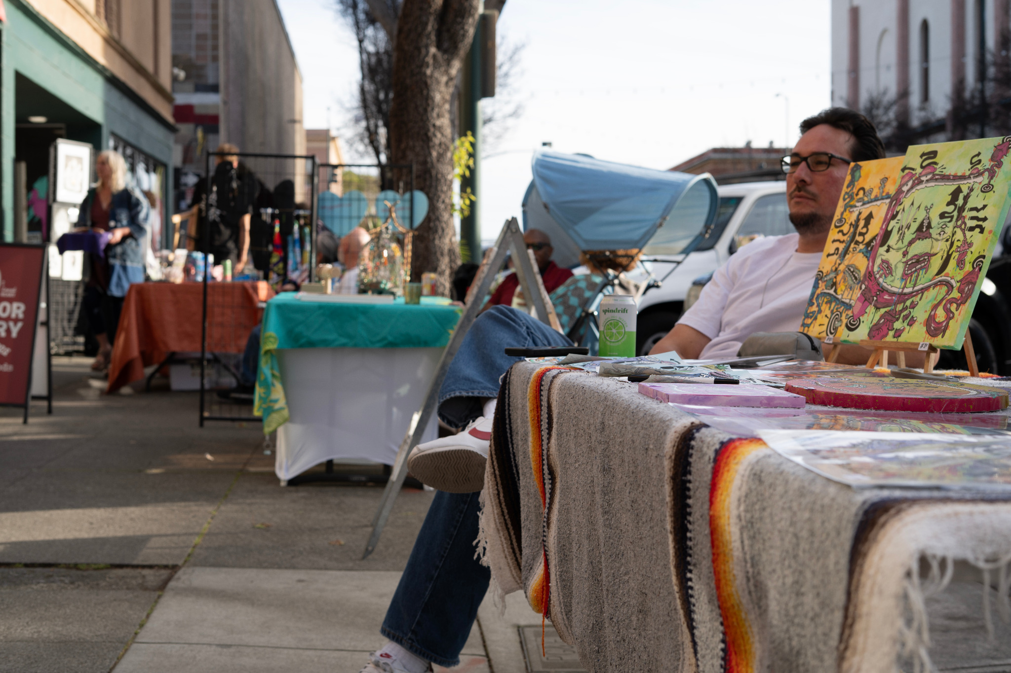 An artist seated behind a display table during an outdoor event.